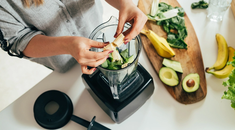 A woman preparing a smoothie with bananas, leafy green, and avocado